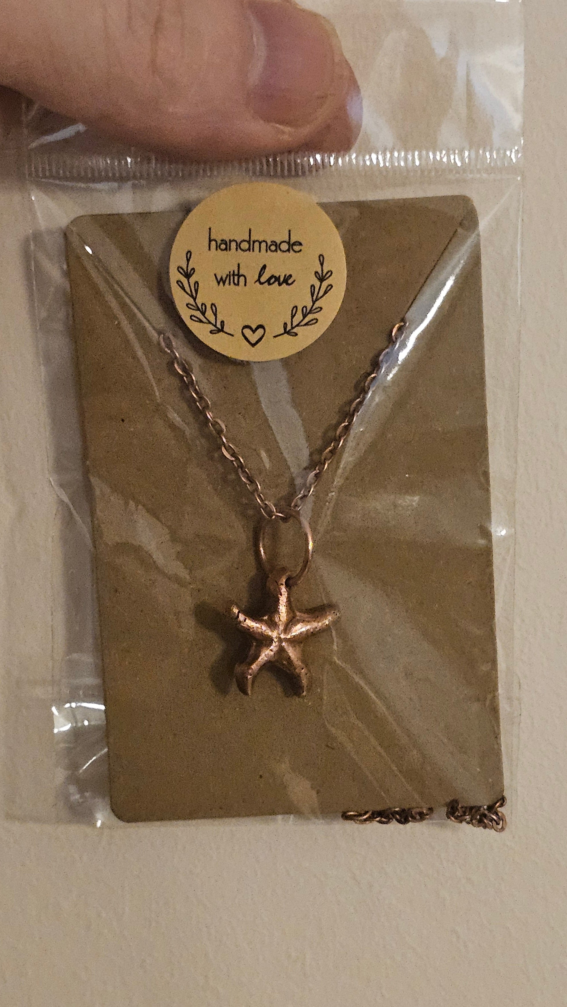 A kiln-sintered copper starfish pendant necklace on a matching chain, displayed on a brown kraft card inside a clear plastic bag with a round 'handmade with love' sticker. A hand is holding the left side of the packaging against a plain beige wall.