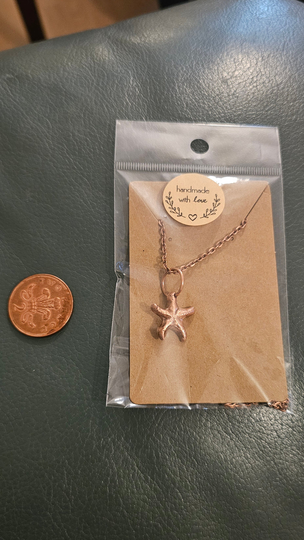 A small kiln sintered copper starfish pendant necklace on a chain, displayed on a kraft paper card inside a clear plastic bag, with a penny below for scale, set on a dark green textured leather surface.