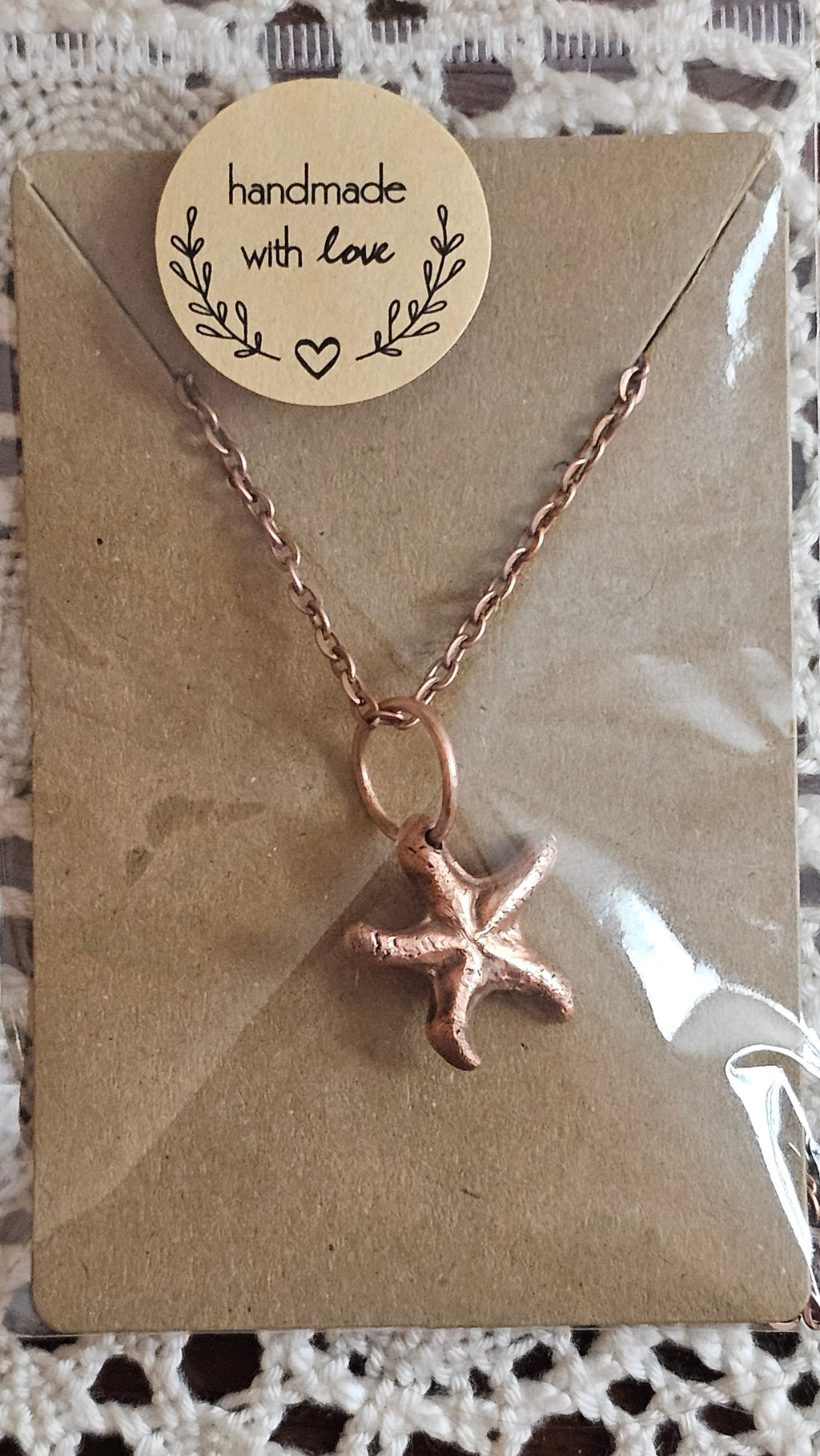 A kiln sintered copper starfish pendant necklace on a delicate chain, presented on a brown kraft paper card with a round 'handmade with love' sticker, wrapped in clear plastic on a white lace background.