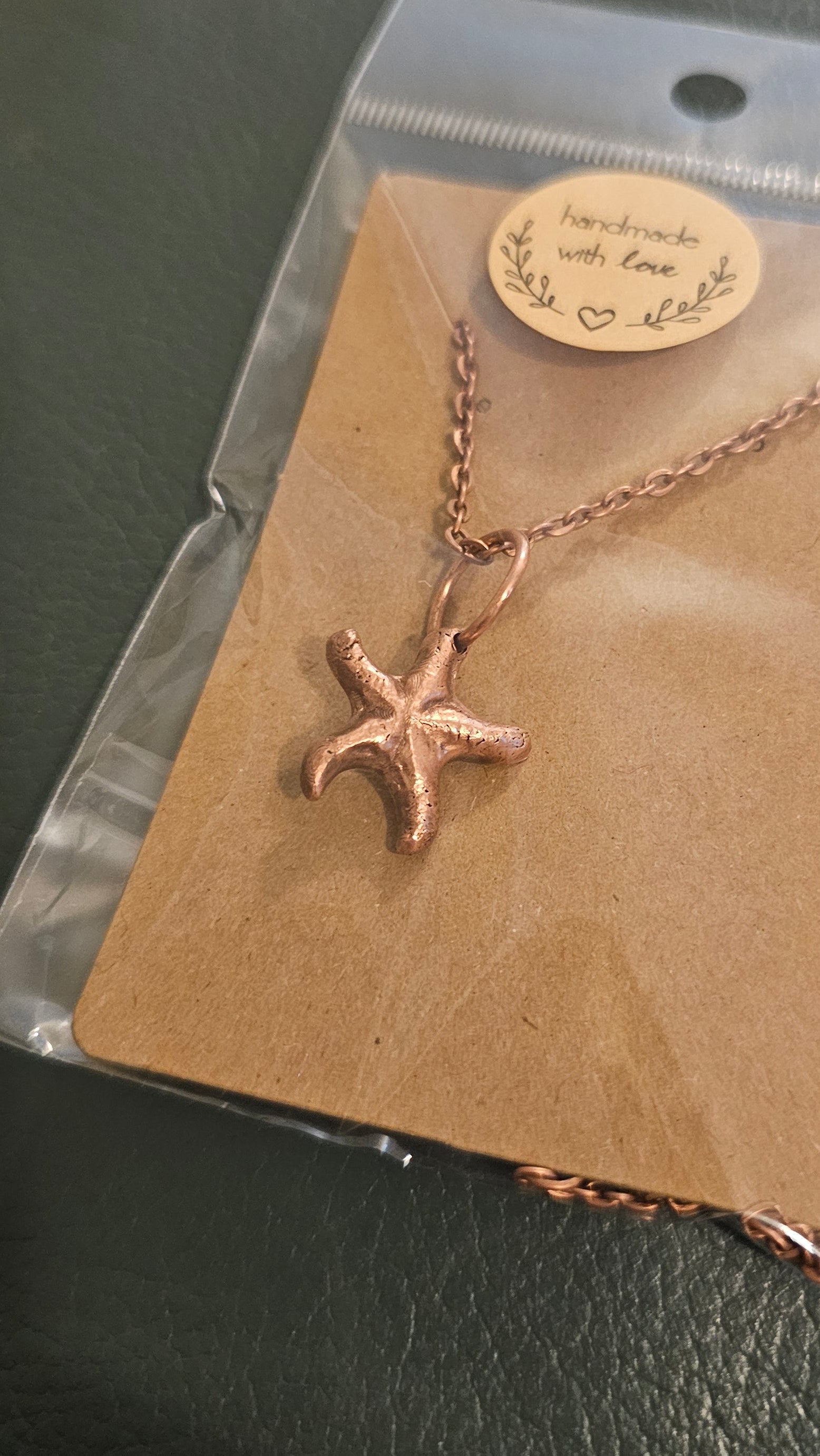 A kiln sintered copper starfish pendant necklace on a matching chain, displayed on a brown kraft card inside a clear plastic bag with a round brand tag, set against a dark textured background.