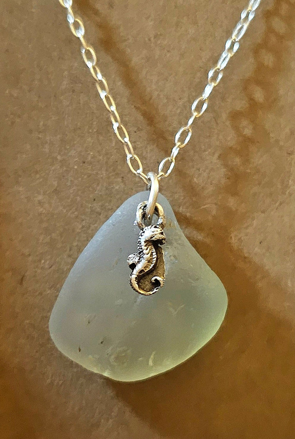 Necklace with an opalescent seaglass pendant and fine silver charm on a sterling silver chain on a beige background as packaged