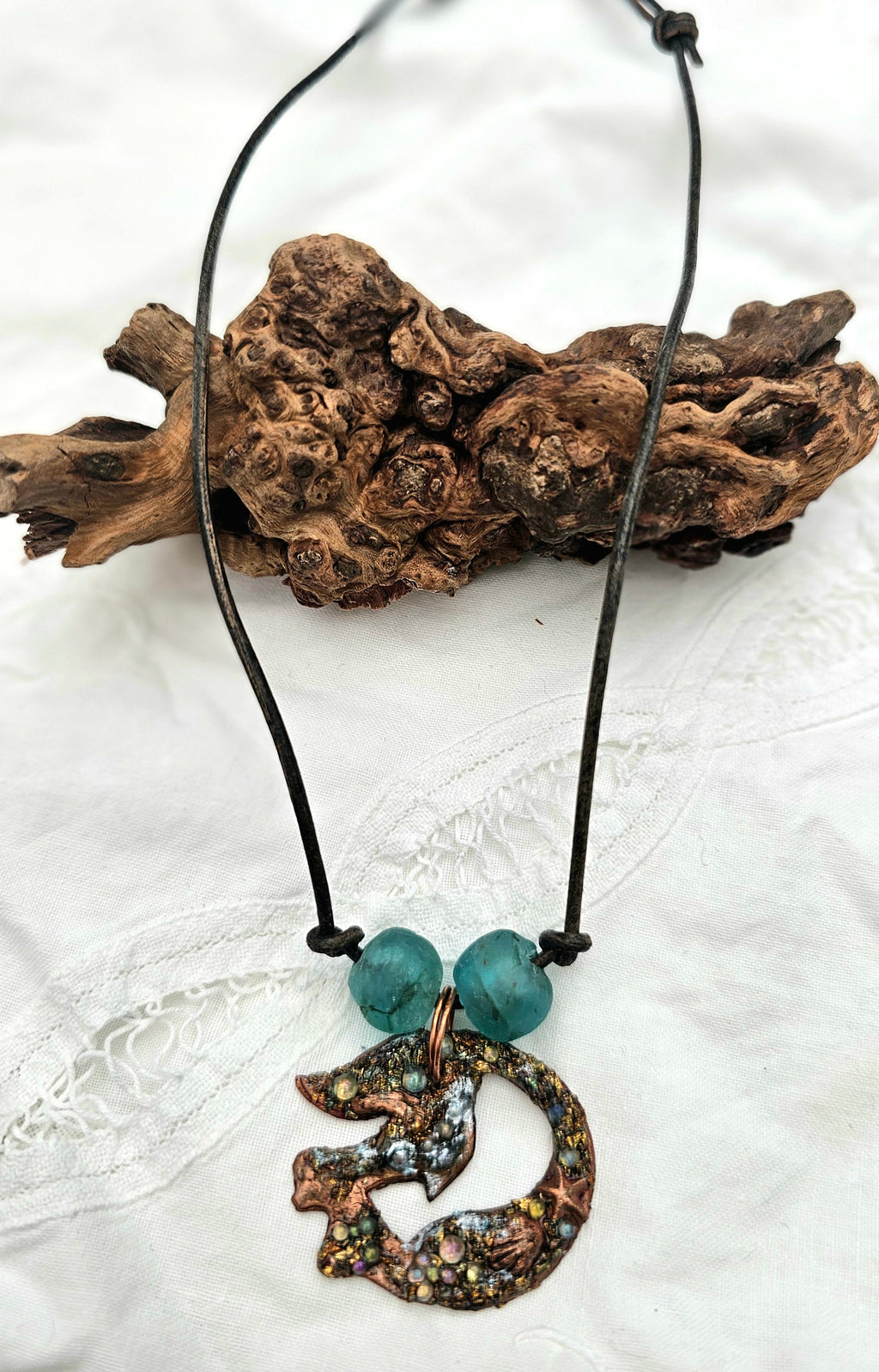 Necklace with a copper mermaid embellished pendant  with glass beads and embellishment on a black leather thong cord, displayed on a beach driftwood background.