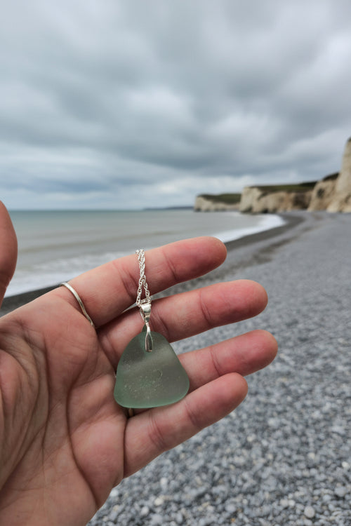 Hand holding a necklace with a seaglass pendant against a neutral coastal background