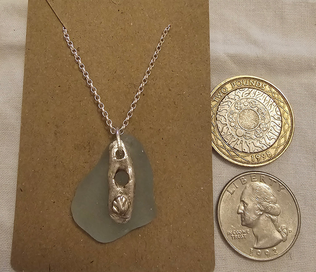 A light blue authentic hand-picked sea glass pendant with fine silver charm and chain with coins for scale.