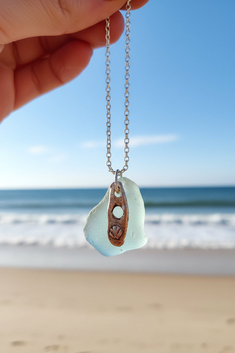 A light blue authentic hand-picked sea glass pendant with fine silver charm and chain against the shore.