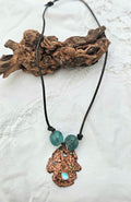 Necklace with a pitted copper pendant and green glass beads draped over driftwood on a white background 