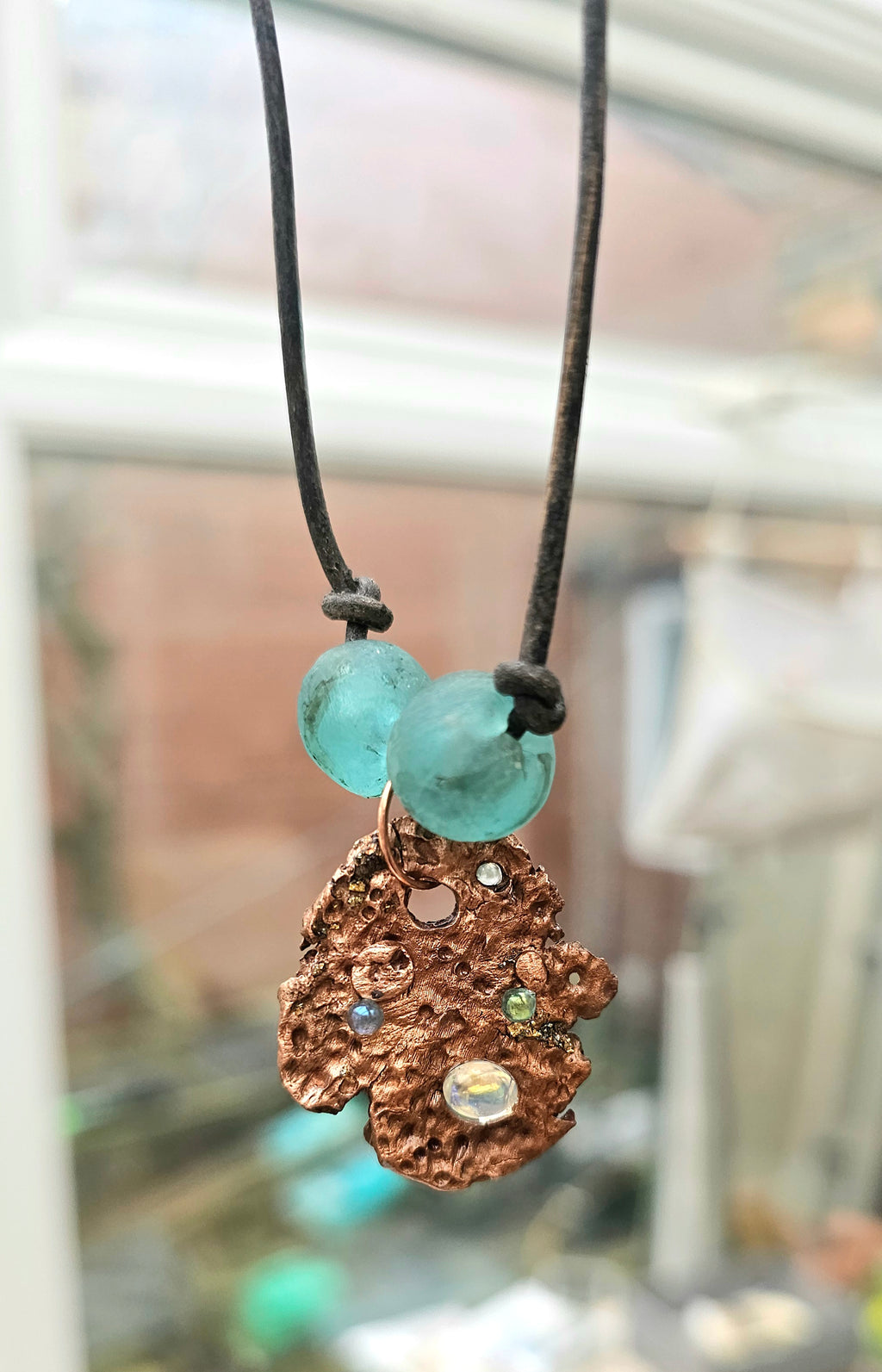Necklace with a pitted copper pendant and turquoise glass beads on a leather thong cord against a blurred indoor background